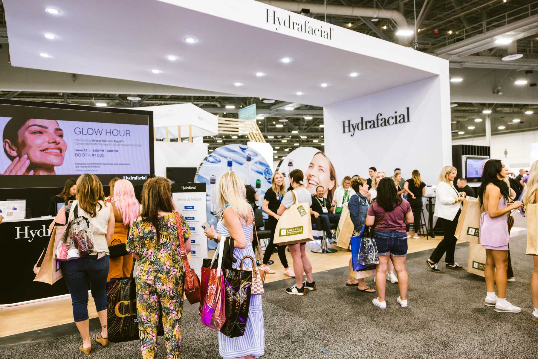 Crowd of attendees gathered in front of a brightly lit Hydrafacial tradeshow booth, with large branded signage, a “Glow Hour” promo screen, and people holding shopping bags and waiting to engage with product demos.
